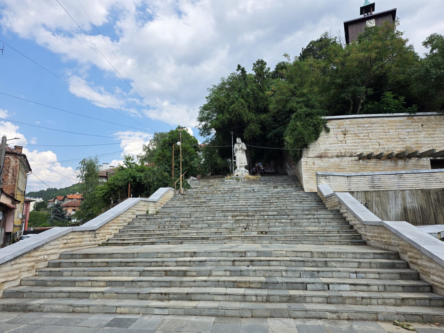 A wide stone stairway with a white statue and a clock tower above it in Dupnitsa