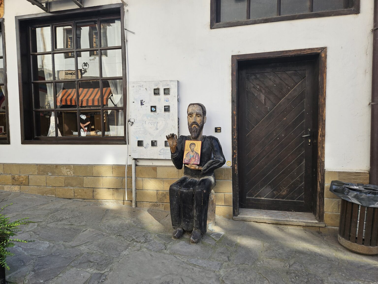 Wooden sculpture of a seated bearded man holding a painted icon beside a dark wooden door on a stone-paved street