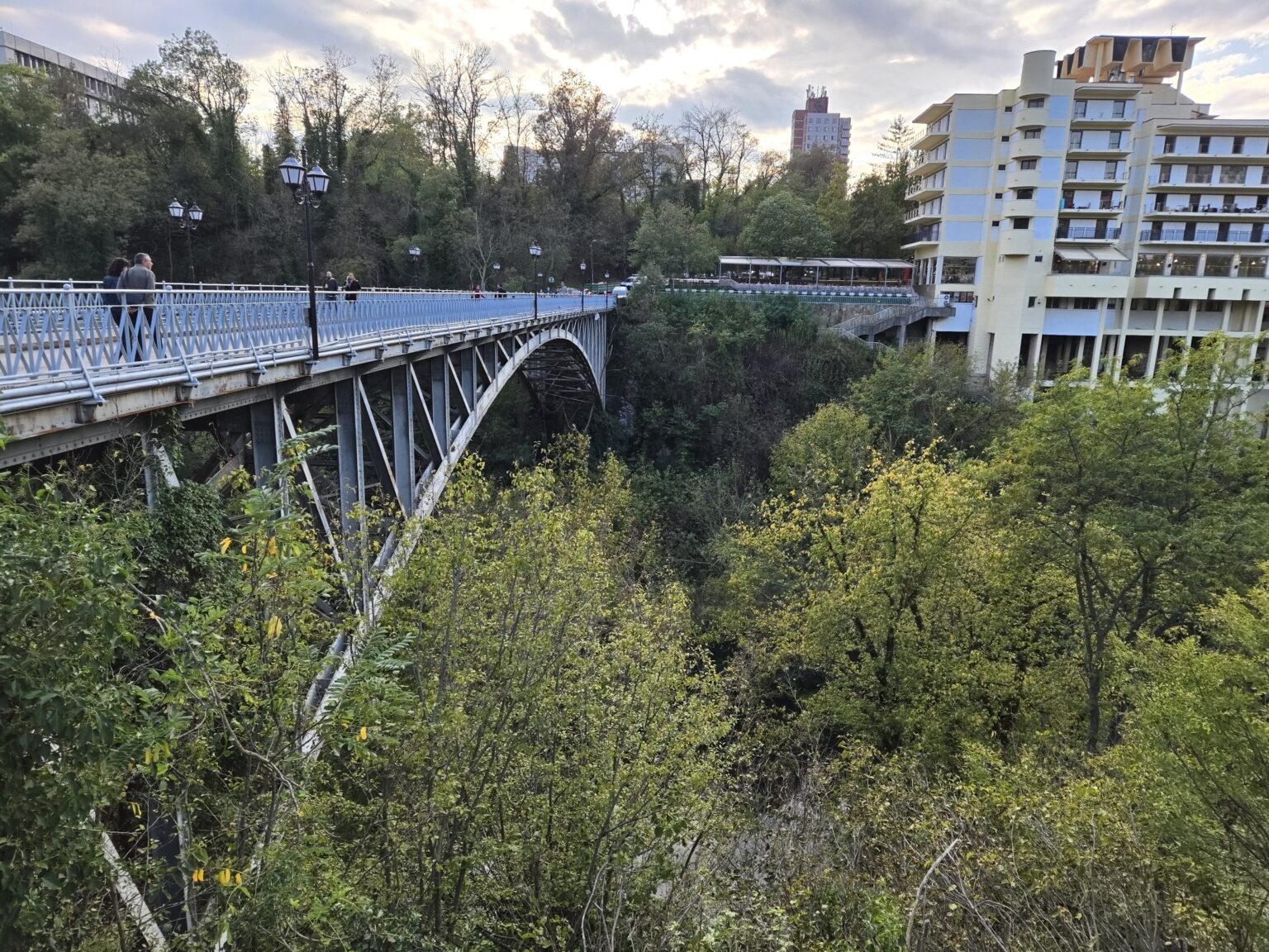Steel arch bridge with pedestrians crossing above a wooded ravine, with a multi-storey building on the right