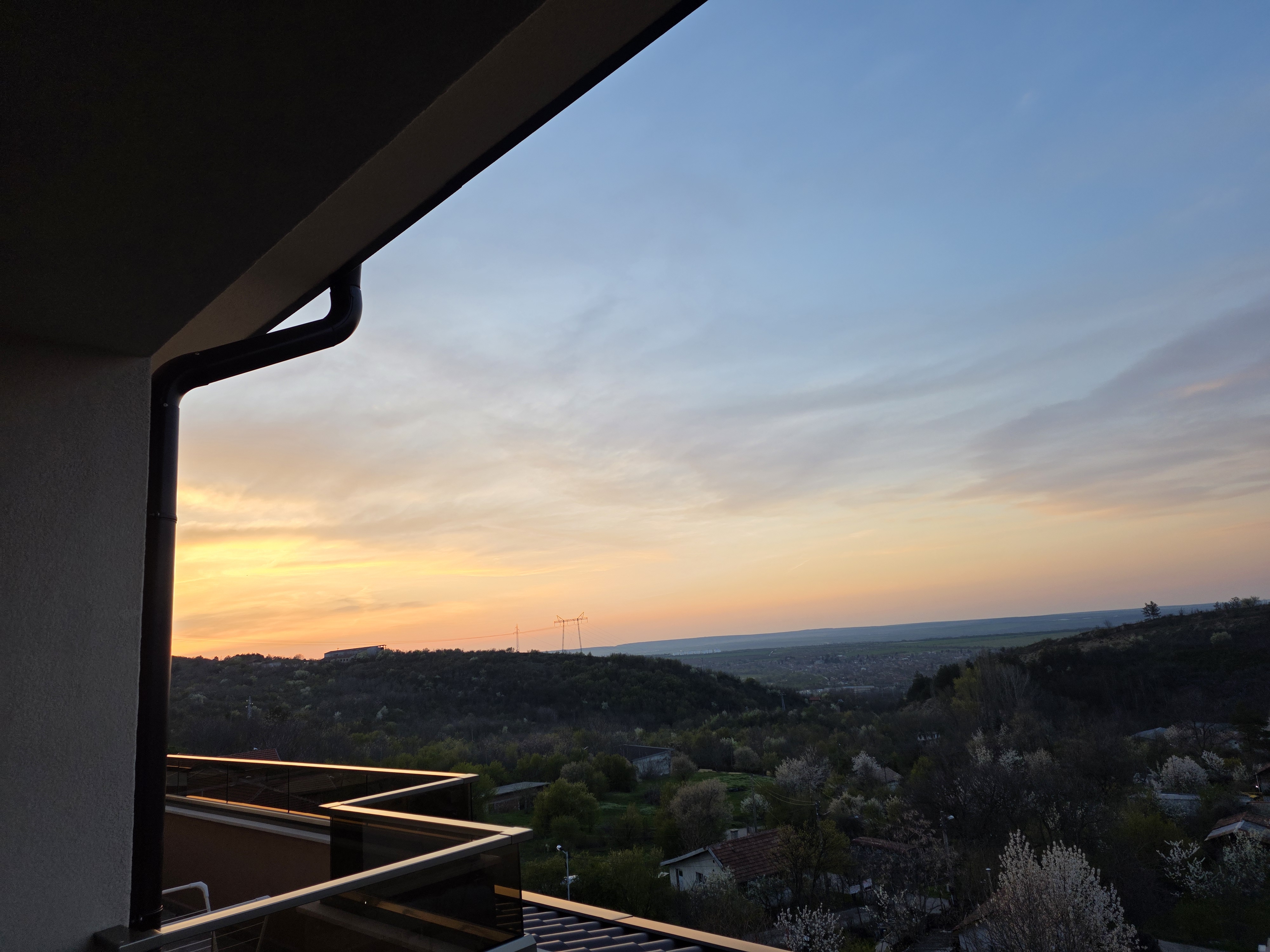 Sunset seen from a terrace in the upper part of Gorna Oryahovitsa, looking over wooded hills, tiled roofs, blossoming trees and power lines above the plain