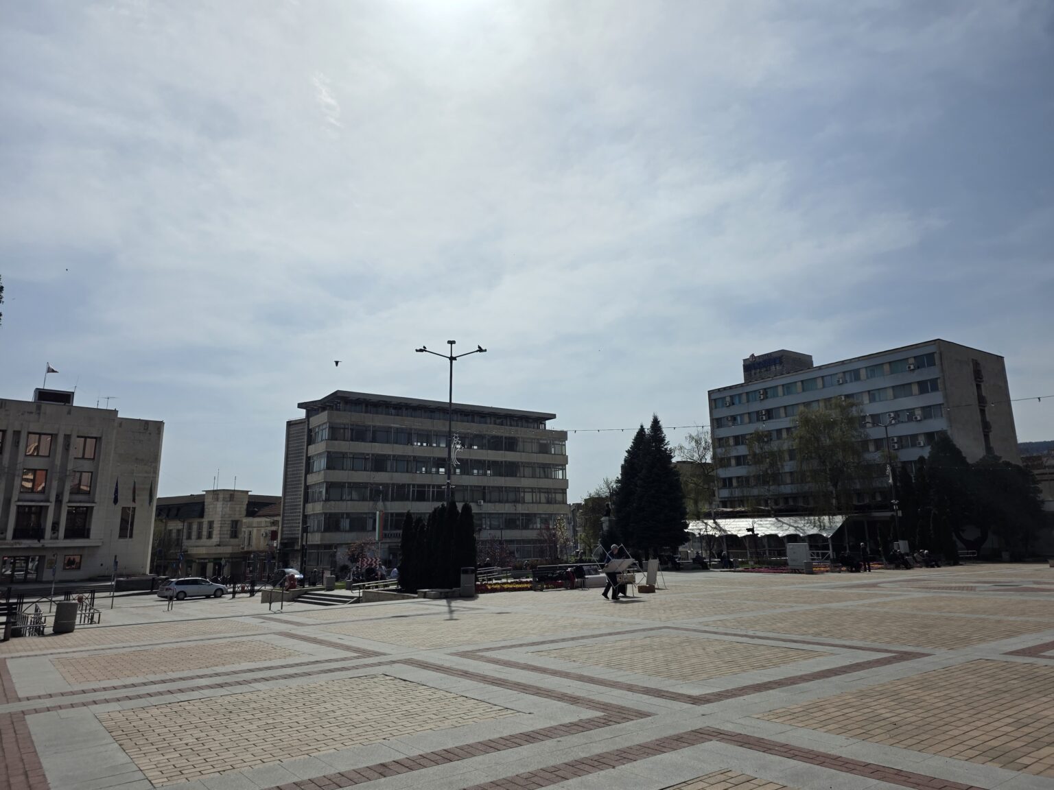 The wide paved main square of Gorna Oryahovitsa with geometric paving, a low administrative building with flags on the left, a glazed office block in the middle and a tall hotel on the right