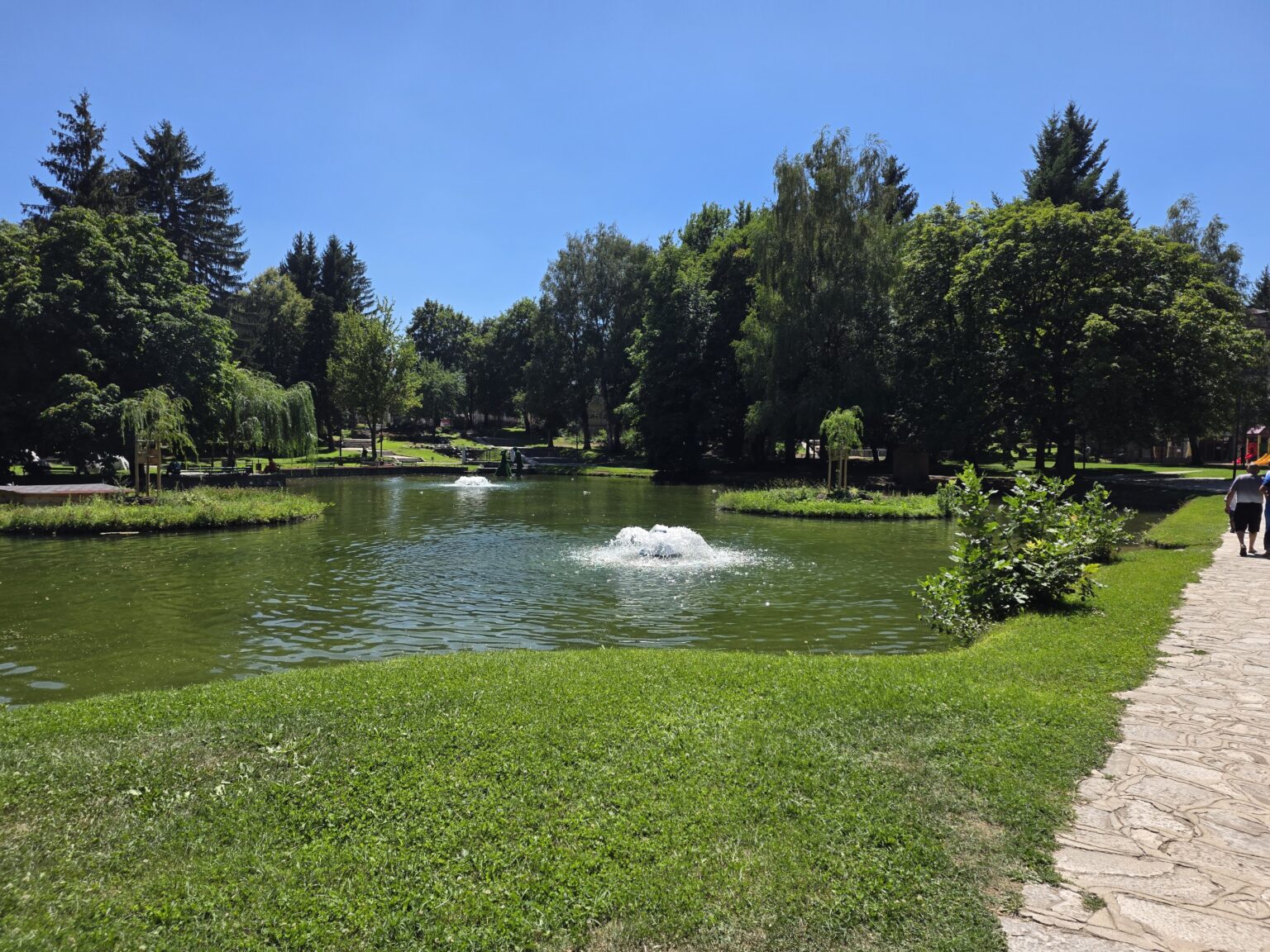 The lake in the park in Razlog with two working water fountains, grassy banks, planted islets and a flagstone path on the right where two people are walking