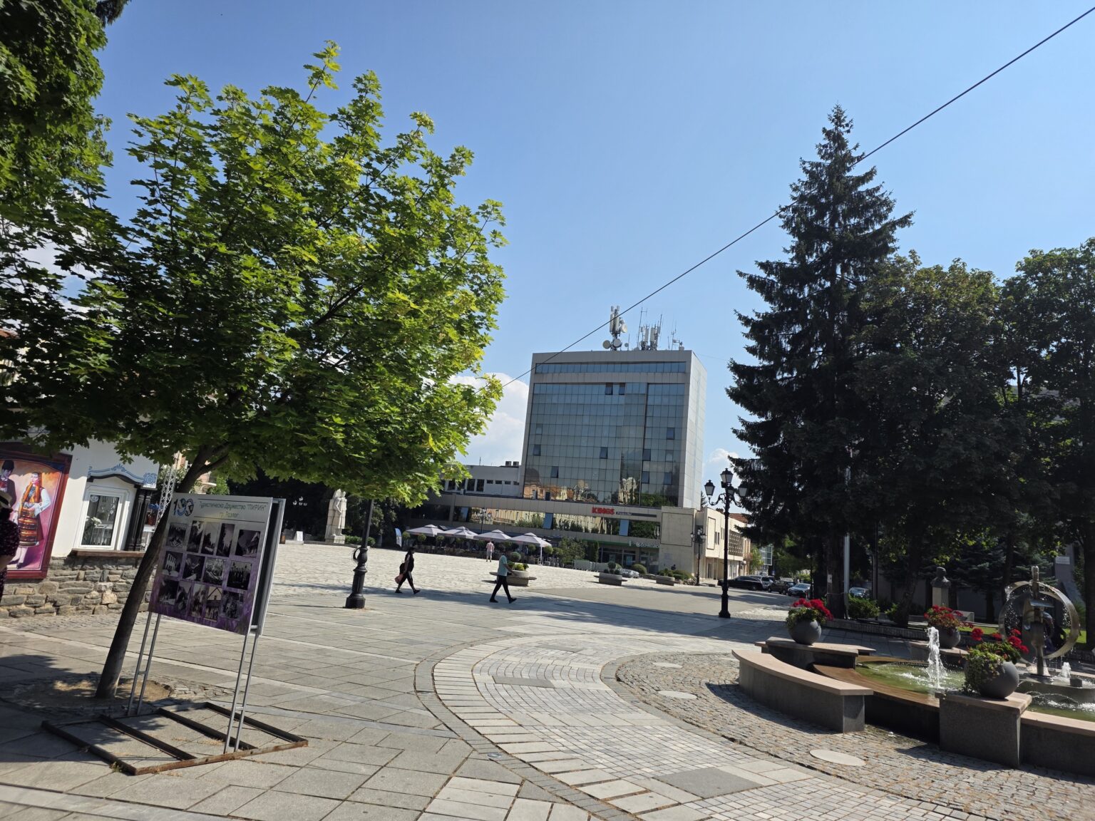 The main square of Razlog with granite paving and a curved cobbled band, a glass-clad office tower in the background, a fountain on the right and a photo exhibition board of the Pirin tourist society on the left