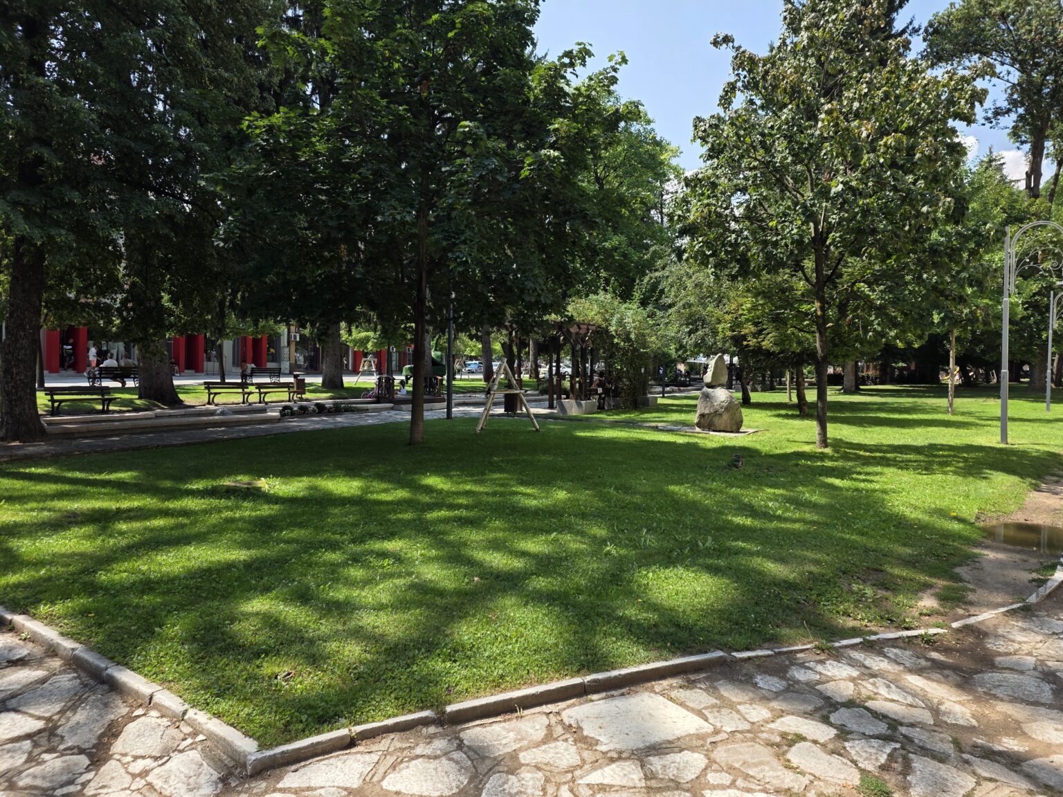 A shaded lawn in the park in Razlog with benches along a stepped path, tall trees, a wooden pergola, a children's climbing frame and a large boulder sculpture on the right