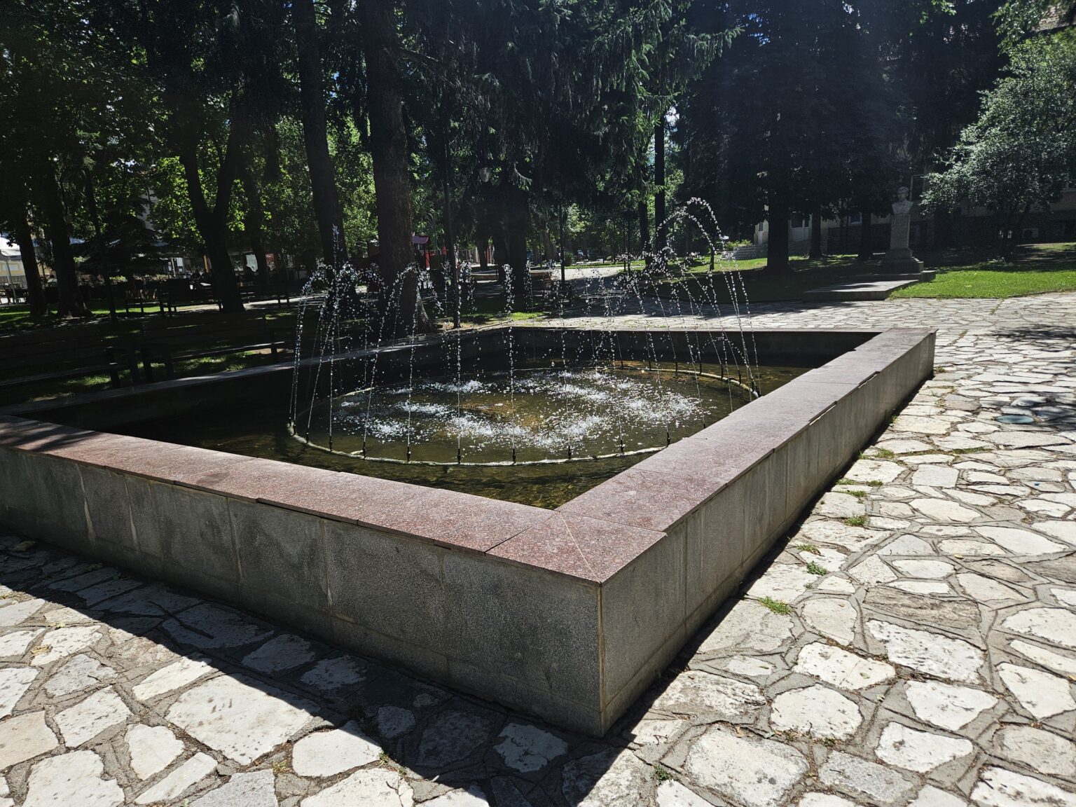 A rectangular fountain with a granite rim and arcing water jets in the shade of the park in Razlog, surrounded by flagstone paving, benches and a bust in the background right