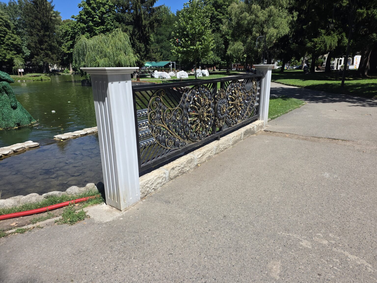 A bridge over the lake in Razlog with white fluted columns and a black and gold openwork railing of wrought rosettes, with shallow water, a stone kerb and a green topiary figure on the left
