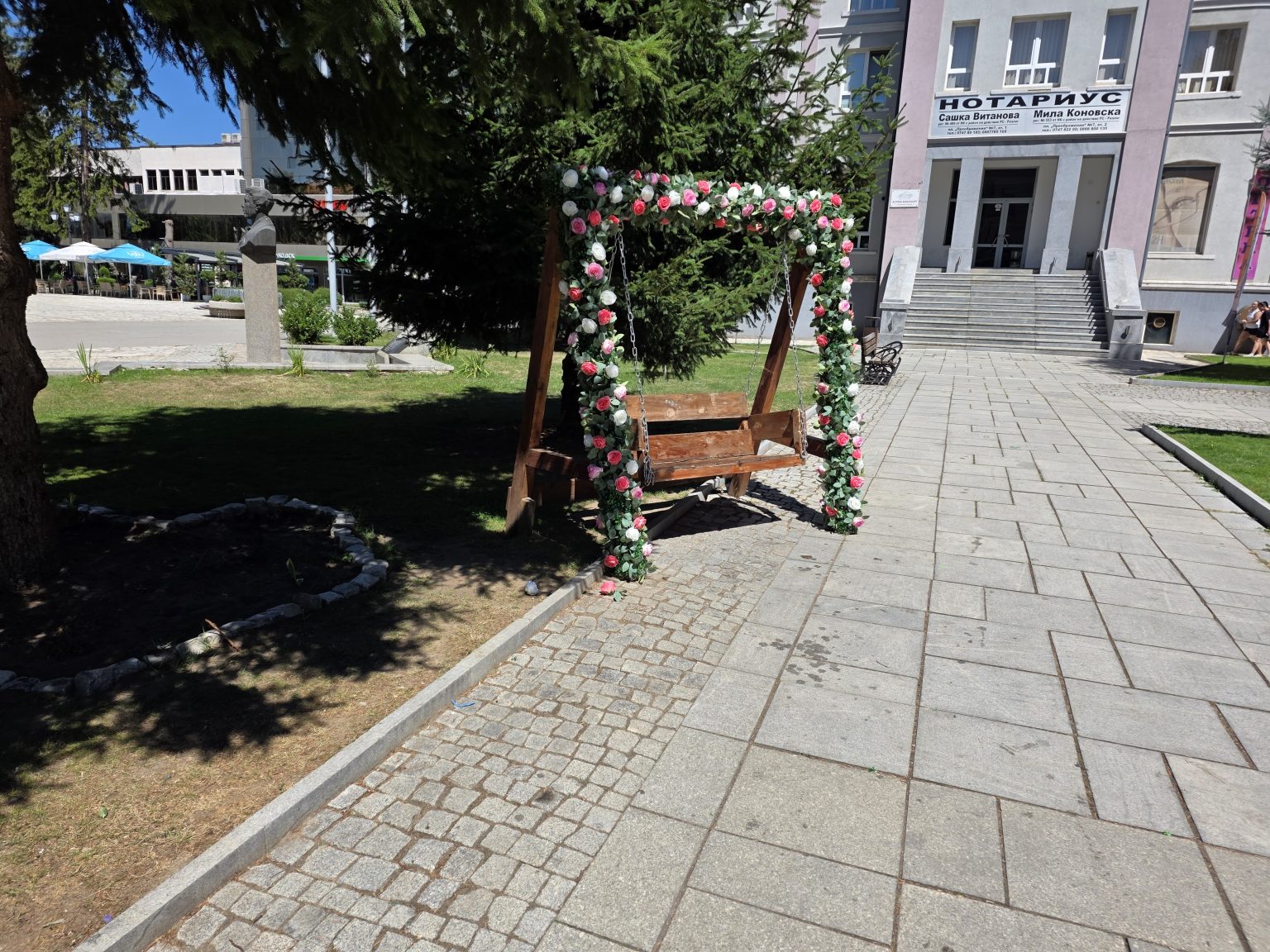 Дървена люлка с рамка, обвита в изкуствени розови и бели рози, до бюст върху гранитен постамент и иглолистно дърво в Разлог, вдясно сграда с надпис Нотариус