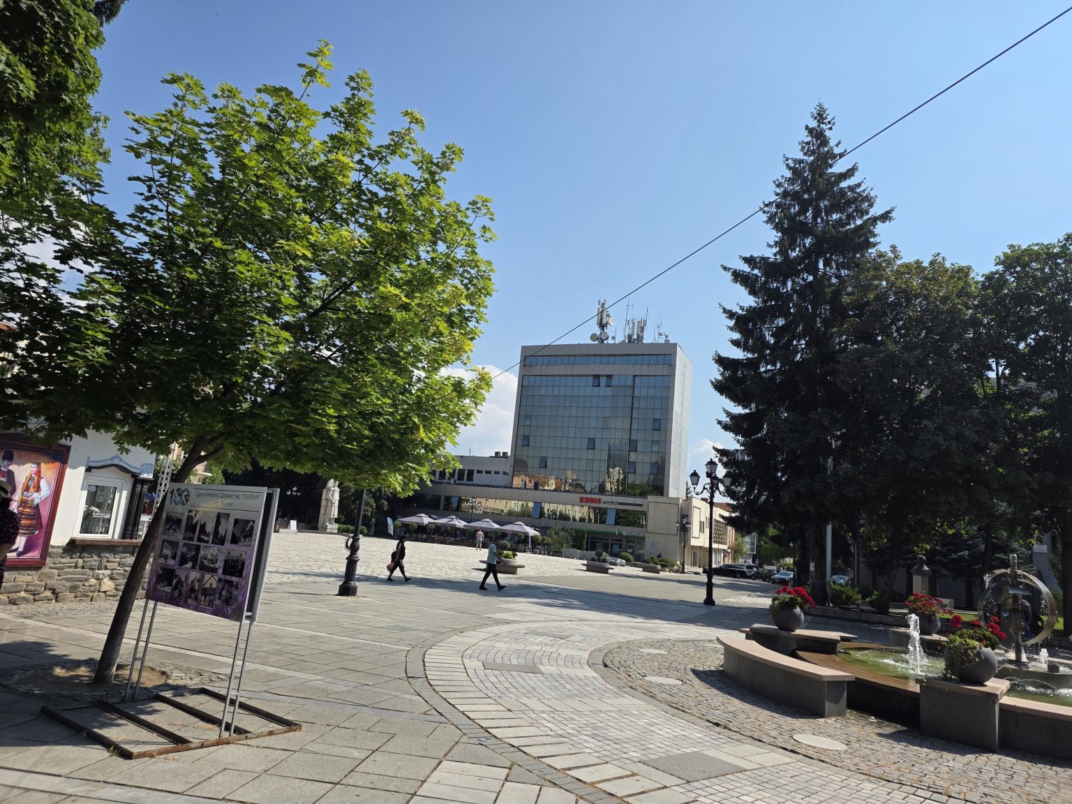 Централният площад на Разлог с гранитна настилка и дъгообразен паваж, остъклена административна кула в дъното, фонтан вдясно и табло с фотоизложба на Туристическо дружество Пирин вляво